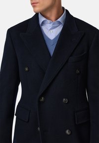 Náměsíční modrý dvouřadý kabát s velkými knoflíky, s klopami a texturovaným povrchem. Převlečený přes světle modrou košili a svetr.