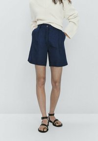 Shorts bleu marine sur mesure avec une coupe décontractée, dotés de poches latérales et d'une fermeture à bouton à l'avant. Portés avec un pull crème et des sandales noires.
