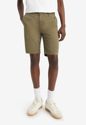 Shorts en coton verts avec une coupe droite, des poches latérales, des passants de ceinture et une texture lisse, associés à des chaussettes blanches et des baskets beiges.