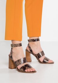 Bruna sandaler med reptilmönster och klack, med två breda remmar och ankelsnörning. Har en kraftig klack som ser ut som trä. Orange byxben synliga.