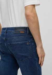 Tmavě modré džíny z denimu s jemnou texturou, s koženou nášivkou označenou nápisem "BOSS" v oranžové barvě a dvěma zadními kapsami.