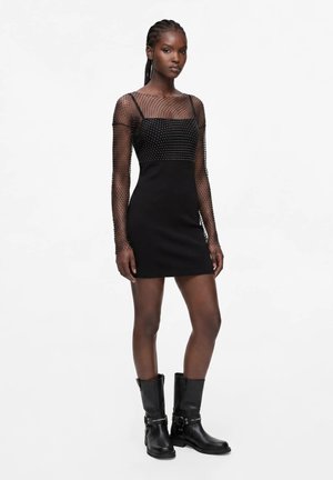 Jeune femme aux cheveux tressés portant une mini-robe noire avec des manches longues transparentes à pois et des bottines noires mi-mollet avec des boucles.