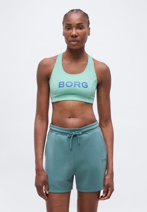 Sprek kvinne som står, iført en mintgrønn sports-BH med teksten "BORG" og turkise shorts med snøring mot en ensfarget lys bakgrunn.