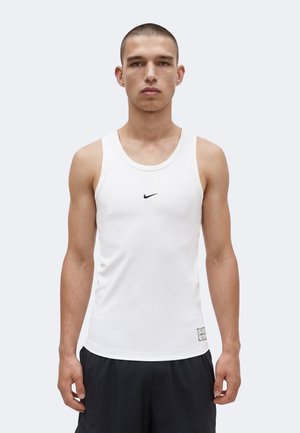 Bărbat tânăr cu păr scurt, purtând un maieu alb Nike fără mâneci și pantaloni scurți negri, stând pe un fundal simplu.