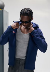 Marineblå hettejakke med glidelås laget av et mykt materiale, med en liten rosa logo, brukt over en grå tanktopp og grå bukser.