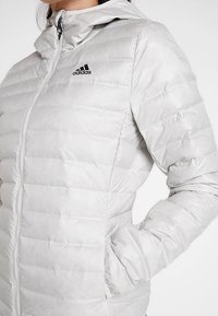 Jasnoszara kurtka ocieplana z kapturem, z zamkiem błyskawicznym z przodu, miękką teksturą i czarnym logo Adidas na górze po lewej stronie.