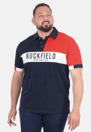 Homme portant un polo du club de rugby français Ruckfield bleu marine et rouge, debout avec un léger sourire et regardant sur le côté.