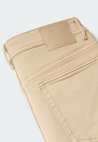 Pantalones de mezclilla beige doblados para mostrar el bolsillo trasero y una etiqueta de cuero marrón claro con la marca "MNG COLLECTION" en la cinturilla.
