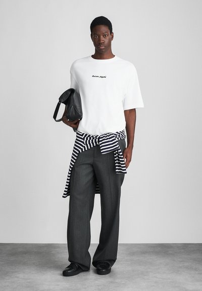 White oversized t-shirt with black text, gray striped shirt tied at the waist, gray wide-leg trousers, and black sneakers, holding a black bag.