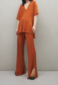 Nainen, joka pukeutuu löysään ribbiverkkopaitaan oranssina ja siihen sopiviin leveisiin housuihin, joiden alla on ruskeat korkokenkäklopit, seisoo yksinkertaista harmaata seinää vasten.