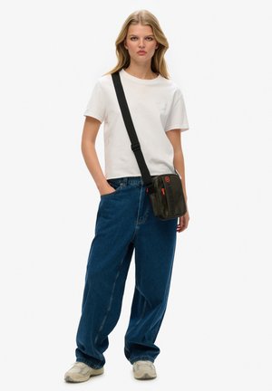 Black crossbody bag with logo, zip closure, and adjustable strap. Model wearing white t-shirt, loose blue jeans, and light sneakers.
