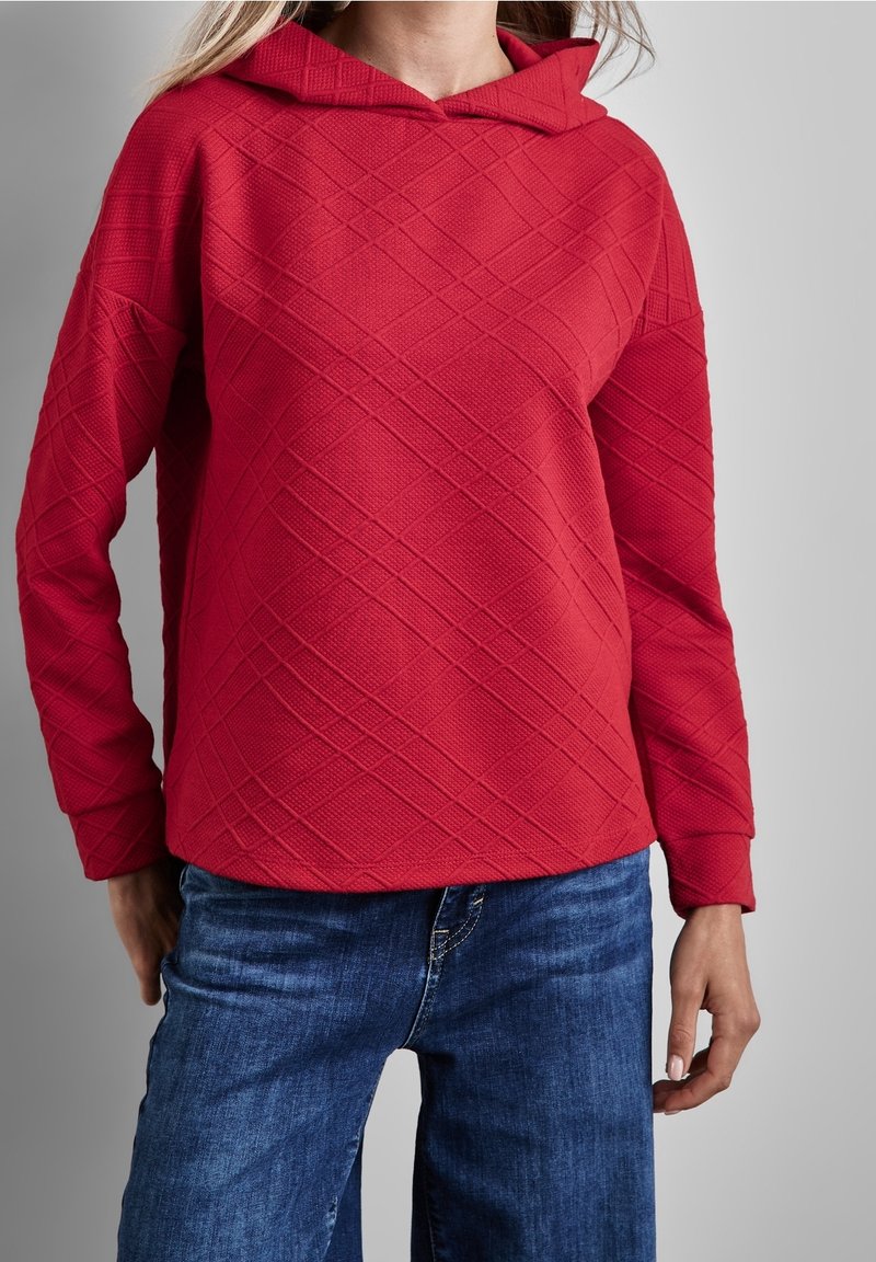 Femme portant un hoodie rouge texturé avec un motif matelassé diagonal et un jean en denim bleu, sur fond gris uni.