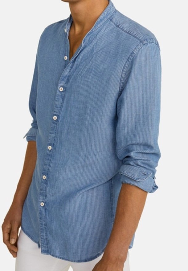 Homme portant une chemise en velours côtelé bleu clair à boutons avec les manches retroussées et un pantalon blanc, debout devant un fond uni.