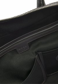 Zwart canvas interieur van een tas met een glad leren afwerking, meerdere vakken en een merklabel met contrasterende stiksels.