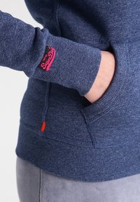 La mano di una persona parzialmente all'interno della tasca anteriore di un hoodie blu lavorato a maglia con il logo rosa "SuperDry" sul polsino della manica.