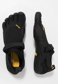 Chaussures noires à cinq orteils avec une tige en maille, sangle ajustable et semelle en caoutchouc. Présente des accents jaunes et des motifs texturés sur la surface.