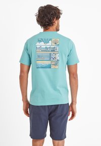 Man facing away wearing turquoise t-shirt with seaside illustrations, including sunset, shell, buildings, waves, puffins, and beach chair.