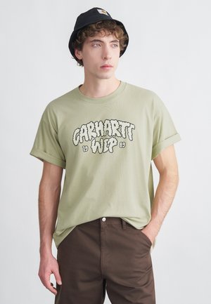 Camiseta de algodón verde claro con mangas remangadas y texto gráfico negro "Carhartt WIP 89". Combinada con pantalones marrones, modelo con un sombrero tipo bucket negro.