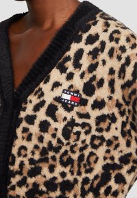 Sweter kardigan w wzór lamparta z czarnymi wykończeniami oraz naszywką z logo Tommy Jeans na piersi, noszony przez osobę o ciemnej karnacji.