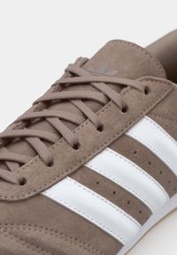Brown suede sneaker with white leather stripes, textured upper, round toe, and flat laces. Features small slots along the sides.
