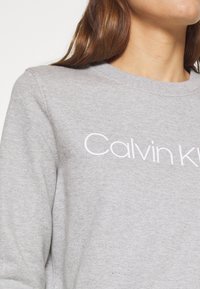 Felpa grigia con collo a giro e logo bianco "Calvin Klein". Il materiale sembra morbido con una texture liscia.
