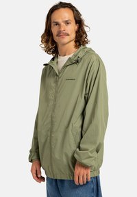 Groene lichtgewicht ritsjack met capuchon, elastische manchetten en twee zijzakken; de stof lijkt glad met een subtiele glans.