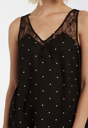 Vrouw draagt zwarte mouwloze top met witte polkadots en kantafwerking rondom de V-hals en schouders.