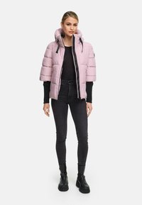 Vrouw in een lichtroze korte mouwen pufferjack met een zwarte lange mouwen shirt, zwarte skinny jeans en zwarte chunky laarzen, staande.