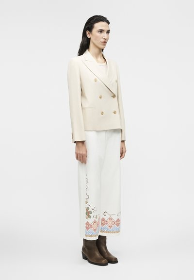 Γυναίκα που φοράει ένα εκρού διπλόμπουκλο σακάκι, λευκό παντελόνι φαρδιάς γραμμής με πολύχρωμη κεντητική κοντά στις άκρες, και καφέ μποτάκια αστραγάλου, στέκεται πλάγια.