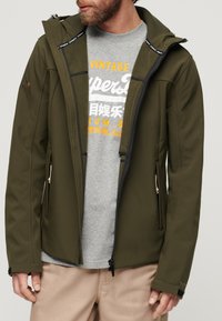 Zielono-oliwkowa kurtka softshell z zamkiem z przodu, regulowanym kapturem i kontrastującymi czarnymi akcentami. Nosi się ją na szarym T-shircie i beżowych spodniach.