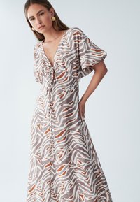 Maxikleidi kerge kangaga, esikinnise ja sügava V-kaelusega, pruunide ja kreemjate abstraktsete lainemustritega. Kergelt puhvis varrukad.