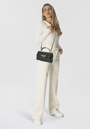 Vrouw in een crèmekleurige geribbelde top en wijde broek met een zwarte, gewatteerde Guess handtas met schouderriem.