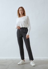 Witte geribbelde trui gecombineerd met donkergrijze taps toelopende broek met een koord, zakken en witte sneakers op een effen achtergrond.