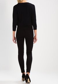 Zwarte slim-fit jeans met een soepele textuur, gecombineerd met een zwart cropped top. Zwarte hoge hakken en een lichte achtergrond maken de look compleet.