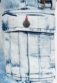 Poche cargo en denim bleu clair avec des coutures visibles, un bouton en argent, et des surpiqûres rouges, présentant une finition texturée et délavée.