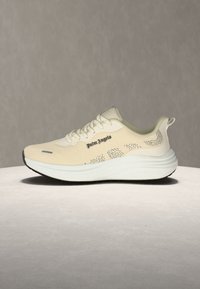 Μπεζ αθλητικό παπούτσι Palm Angels με λευκή παχιά σόλα, μαύρο περιθώριο σόλας και διακριτικό πουά σχέδιο στις πλευρές, παρουσιάζεται σε ουδέτερη επιφάνεια και φόντο.