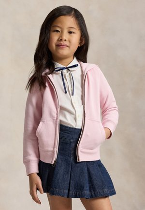 Niña joven con el pelo largo y oscuro, que lleva una camisa blanca con lazo azul marino, sudadera rosa con cremallera y falda azul oscuro plisada, de pie con una mano en el bolsillo.