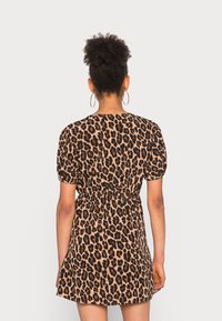 Femme aux cheveux bouclés en chignon haut, portant des boucles d'oreilles créoles dorées et une robe courte à imprimé léopard avec des manches bouffantes, vue de dos.