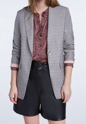 Kvinde iført en grå ternet blazer over en lyserød mønstret bluse og sorte højtaljede lædershorts med knap- og lynlås.