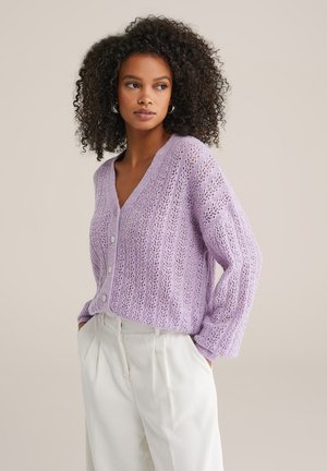Cardigan lavorato a maglia viola con scollo a V, caratterizzato da motivi traforati e bottoni sulla parte anteriore. Abbinato a pantaloni bianchi plissettati.