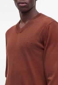 Jeune homme portant un pull marron à col en V avec "BOSS" brodé sur la poitrine gauche, sur un fond uni clair.