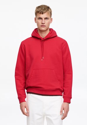 Junger Mann in einem roten Kapuzenpullover mit Fronttasche, stehend vor einem schlichten weißen Hintergrund, trägt weiße Hose.