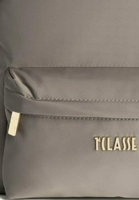 Zaino di tessuto grigio con tasca, zip dorata e logo del brand "1ª CLASSE" in oro sulla parte anteriore.