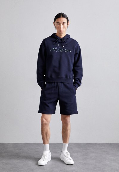 Hoodie azul-marinho com cordão e texto "Missoni" em lantejoulas coloridas, combinado com calções azul-marinho e ténis brancos.