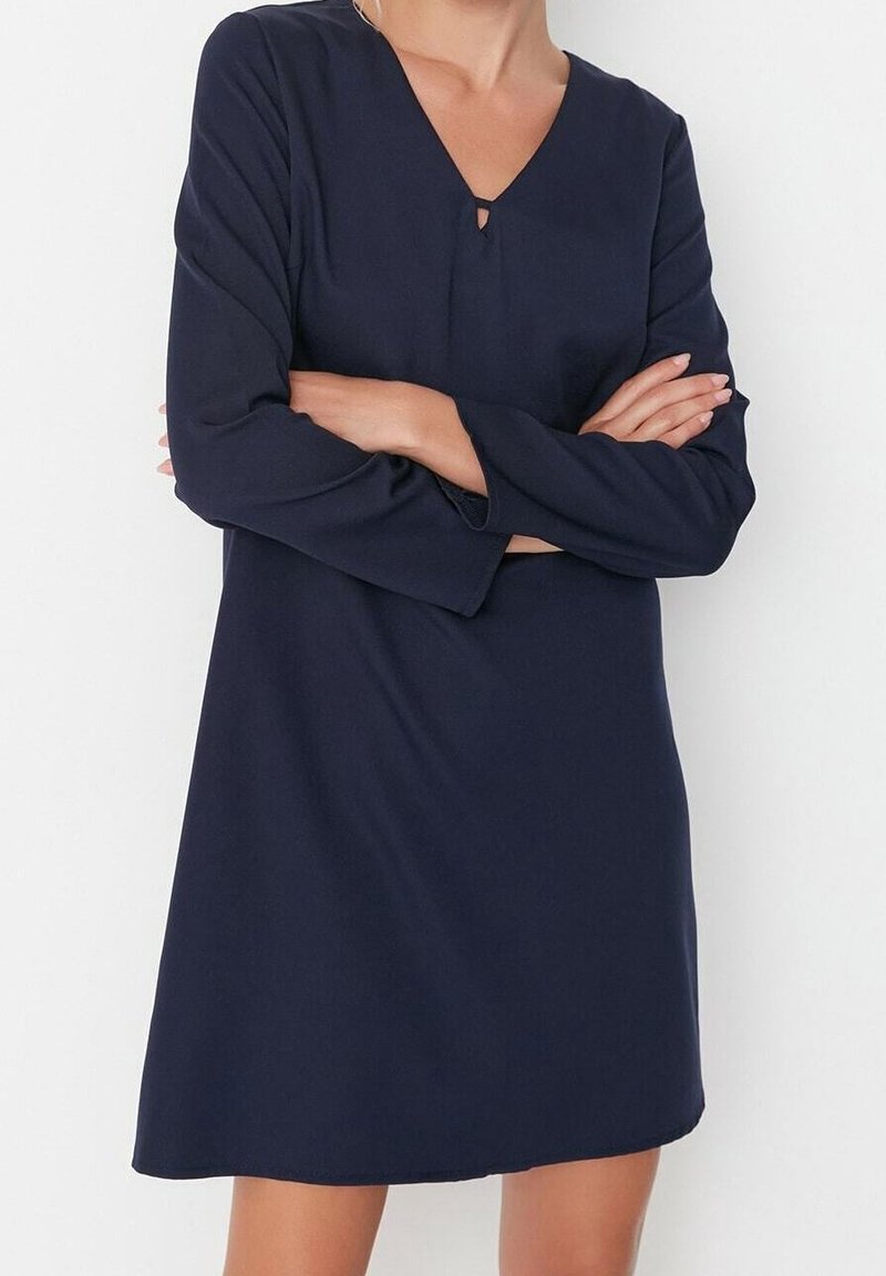 Robe bleu marine avec un décolleté en V, des manches longues et un petit détail en forme de trou à la poitrine. Tissu lisse et léger ; design arrivée au genou.