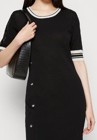 Femme portant une robe en maille noire avec un col et des manches rayés blancs, tenant un sac à bandoulière noir texturé.