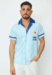 Chemise à manches courtes bleu clair avec un col uni bleu marine, un graphisme floral et le texte "VIVRE LA VIE", présentant des accents rayés bleus sur les manches.