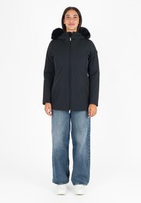 Zwarte geïsoleerde jas met een capuchon met bontrand, ritssluiting, zijzakken en gecombineerd met losse blauwe jeans en witte sneakers.