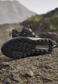Dark green hiking boots with rugged tread soles resting on rocky ground outdoors with blurred mountains in the background.