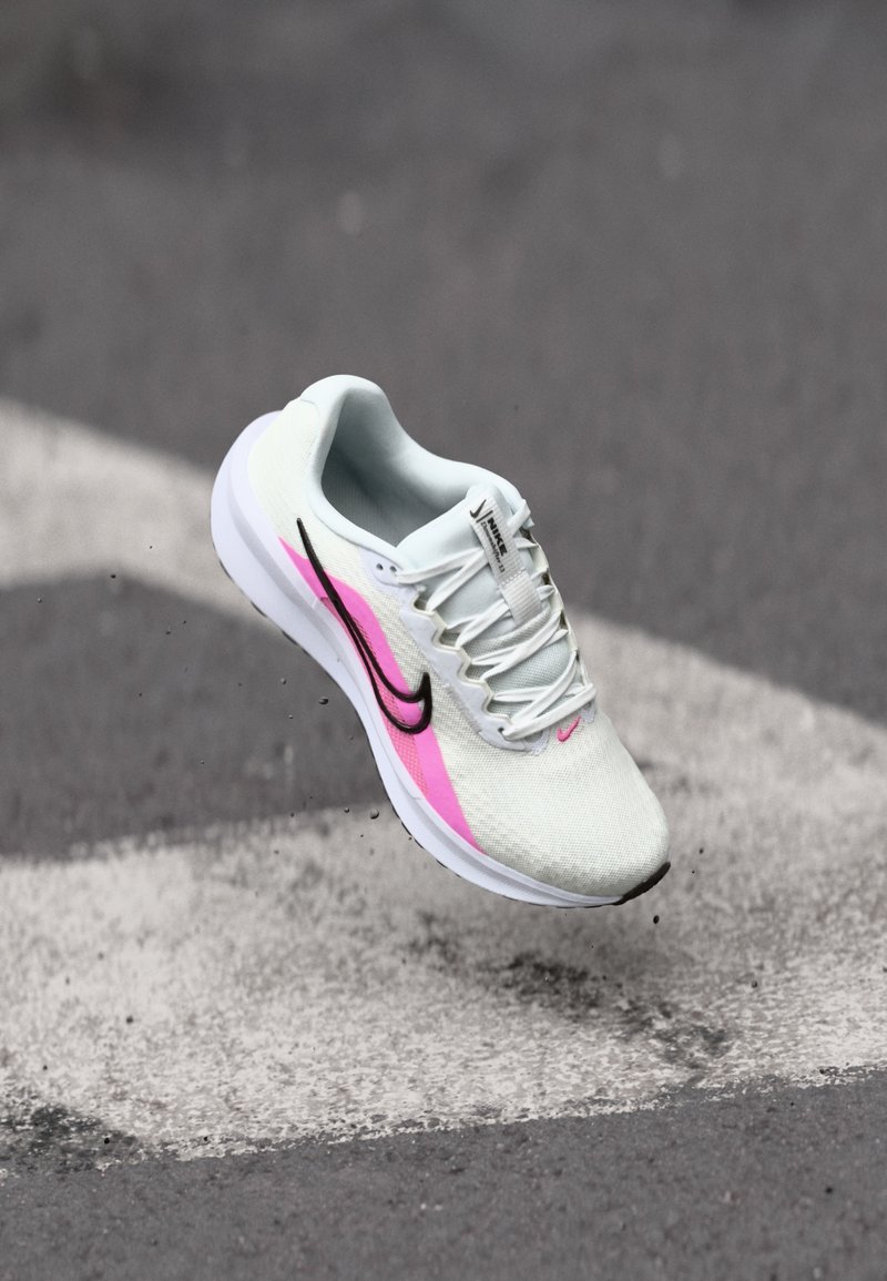 Chaussure de course légère en maille blanche, avec des accents roses et noirs, talon surélevé et semelle texturée, positionnée en l'air au-dessus du pavé.